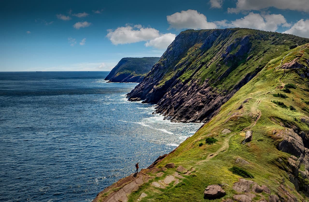 A person stands on a grassy cliff overlooking the ocean, with steep rocky cliffs and a distant horizon under a partly cloudy sky.